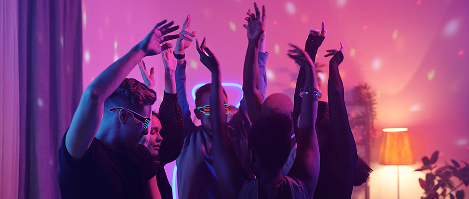 Νεαροί διαπολιτισμικοί φίλοι με έξυπνα καθημερινά ρούχα σηκώνοντας τα χέρια ενώ χορεύουν μαζί στο πάρτι στο σπίτι στο σαλόνι φωτισμένο με ροζ φωτισμό